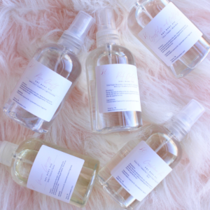 Ariel view of five dry body oils in clear four ounce spray bottles laying on a pink fur pillow