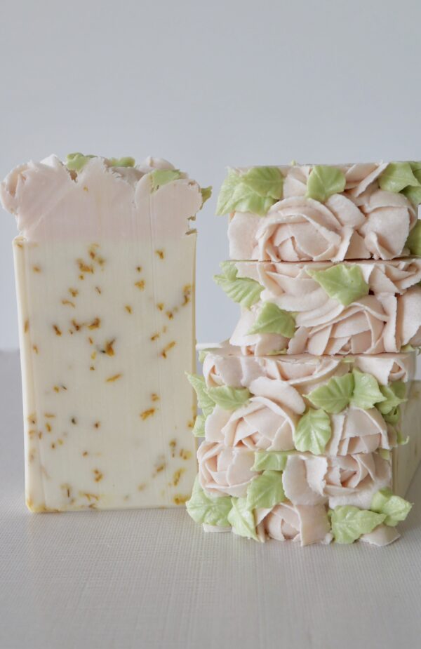 A stack of lemon rosewater bars with lots of pink piped soap roses on top and a cut bar facing front showing calendula petals inside