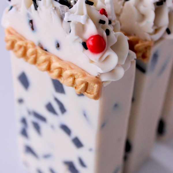 Close up view of two Cruella Couture Bars with cream base and black chunks scattered throughtout. A gold soap chain at the top of the base of the bar and piped white soap with black and red sprinkles on top