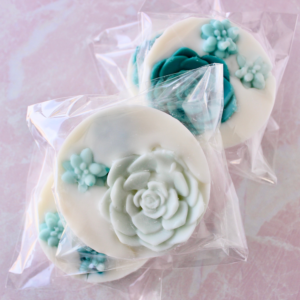 Three stacked round wax melts with succulent wax embeds inside that are wrapped in clear bags. Pink Marble background.
